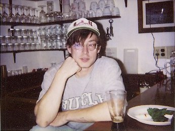 Kevin Blisske at Palmer Station in 1994 — ball cap, wire-frame glasses and an oversized sweatshirt, chin propped on one hand at the station bar, a drink and a plate in front of him.