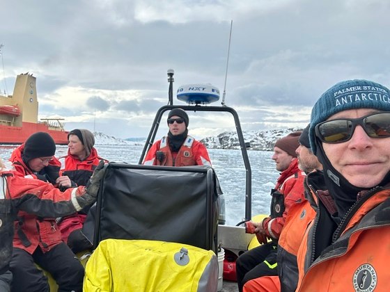 Kevin Blisske at Palmer Station in the 2025-26 season, front right in a Zodiac inflatable with six colleagues in orange float coats. He wears wraparound sunglasses and a beanie reading PALMER STATION ANTARCTICA. Behind them: the orange-hulled research vessel Laurence M. Gould, sea ice and a glacier.